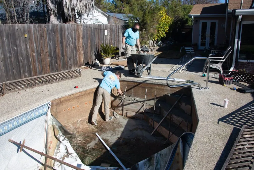 Two workers maintaining a drained backyard pool during renovation, one standing in pool basin and one operating equipment nearby