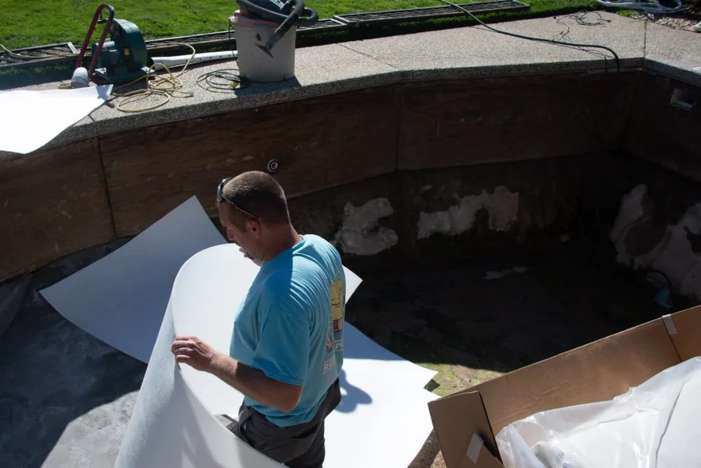 Worker installing white pool liner into excavated residential pool with equipment visible at edge