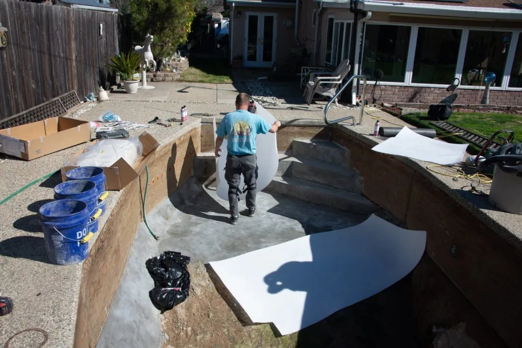 Worker standing in backyard pool construction area with concrete forms, buckets, and building materials