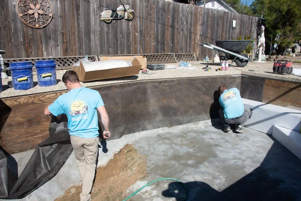 Two workers in blue shirts preparing concrete pool construction in an excavated pit surrounded by wooden fencing and equipment.