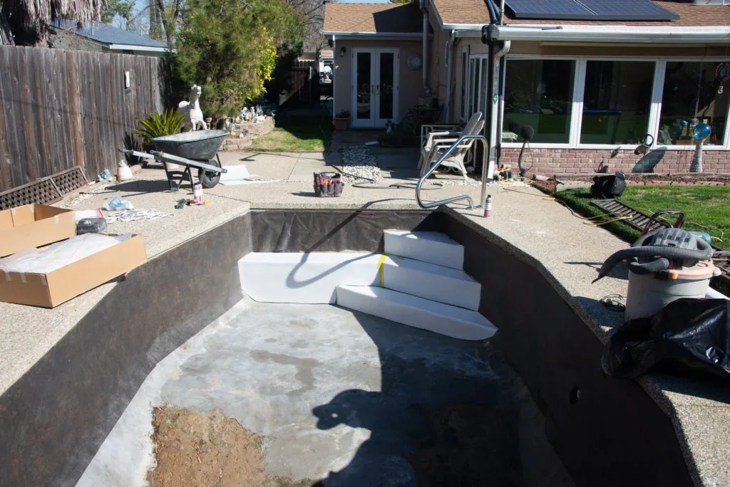 In-ground pool under construction with concrete walls, white steps, and construction materials scattered in backyard of brick home