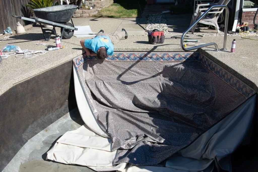 Person in blue shirt cleaning pool with hose and textile liner in sunny backyard setting