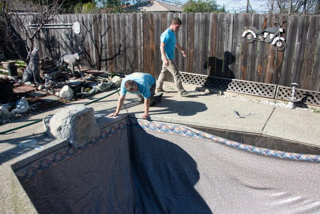 Two people installing pool liner in backyard with wooden fence and maintenance equipment visible