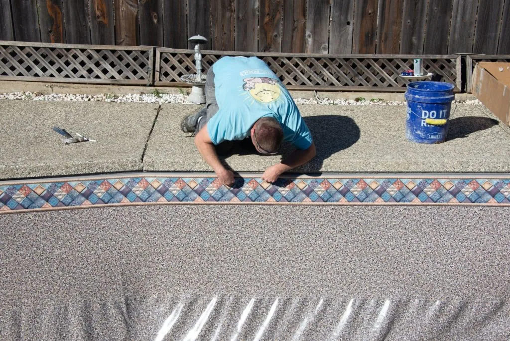 Worker in blue shirt installing decorative tile border along pool deck next to wooden fence