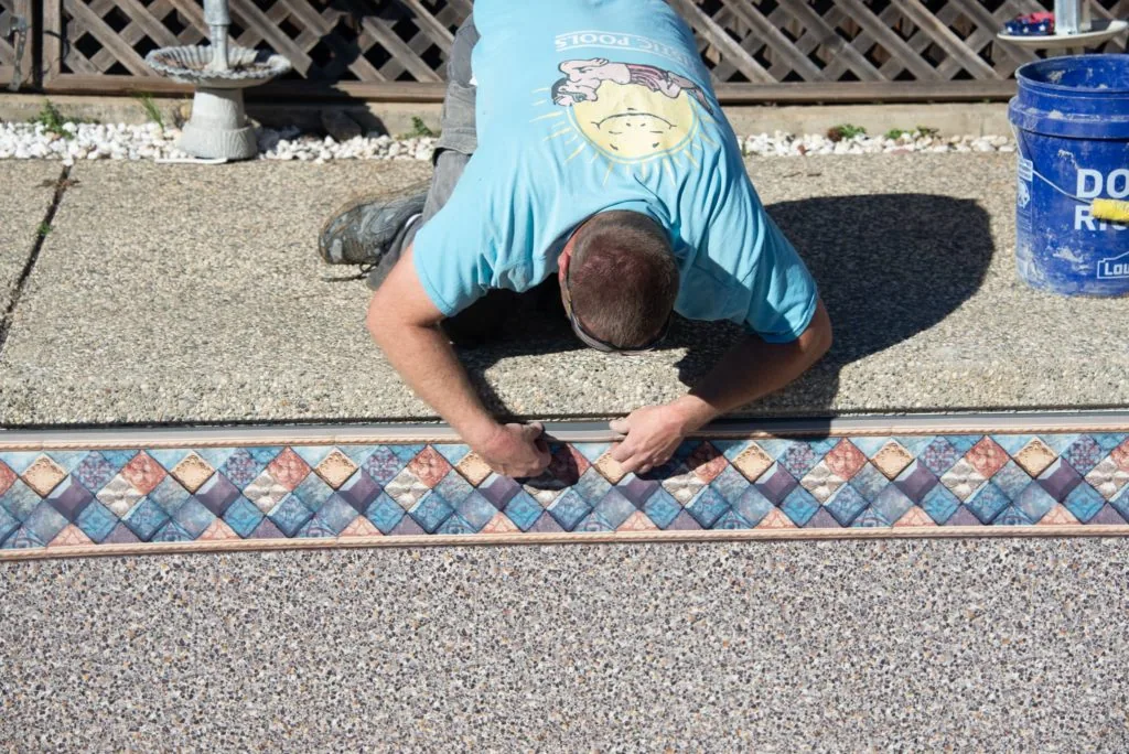 Person in blue shirt installing decorative tile border along pool edge