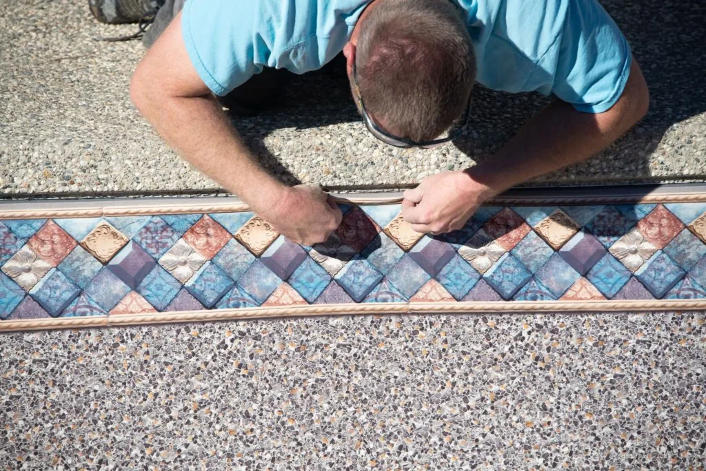 Man in light blue shirt installing decorative tile border along pool deck edge