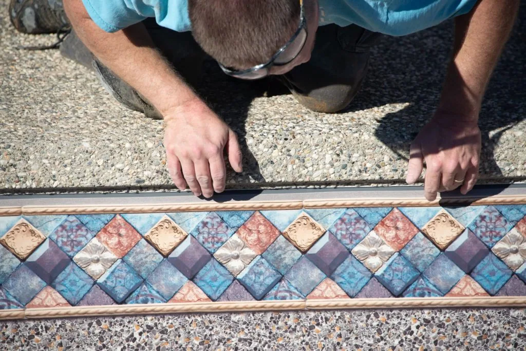 Person's hands positioning decorative tile border with diamond pattern and blue, coral, and cream colors against concrete surface
