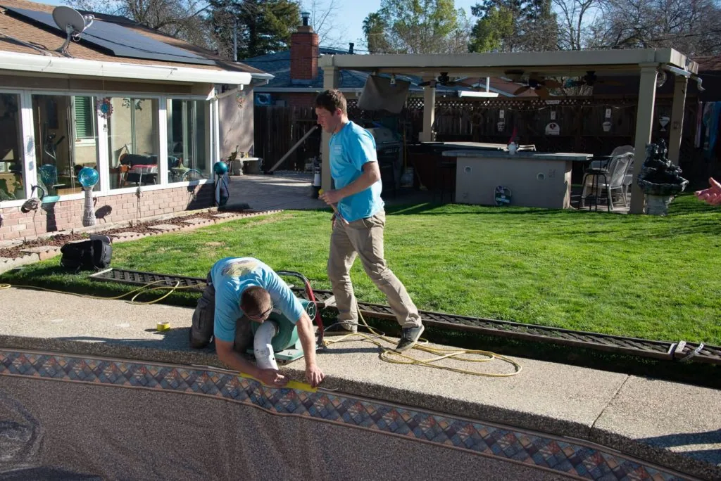 Two people installing patterned material along the edge of a backyard pool during daytime construction