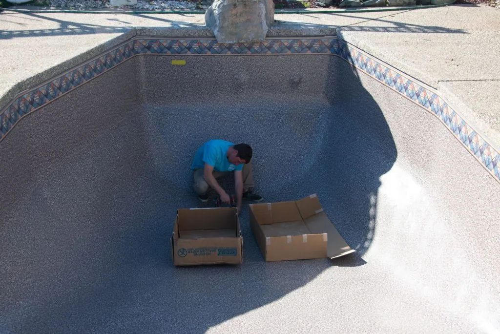 Person in blue shirt crouching in empty pool with cardboard boxes
