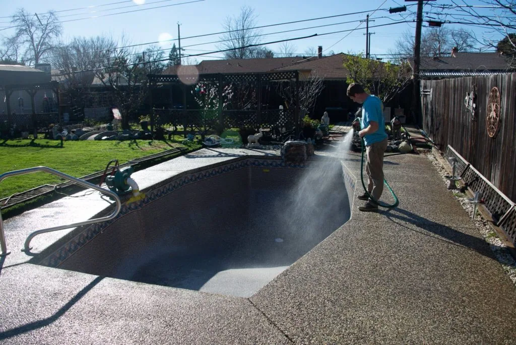 Person pressure washing a concrete pool deck in a residential backyard with an empty pool visible
