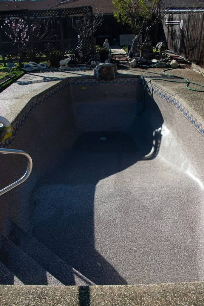 Empty residential pool with dark interior and concrete deck surrounded by landscaping and black fence