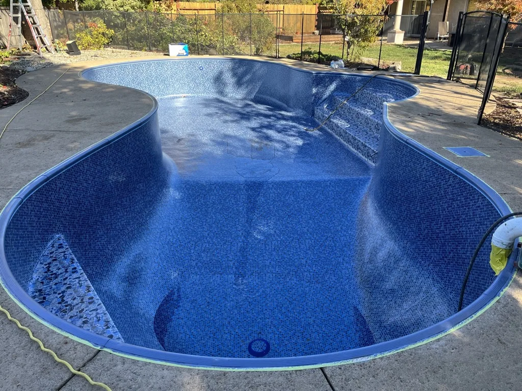 Empty backyard swimming pool with blue tile interior, concrete deck, and metal fence surrounding it