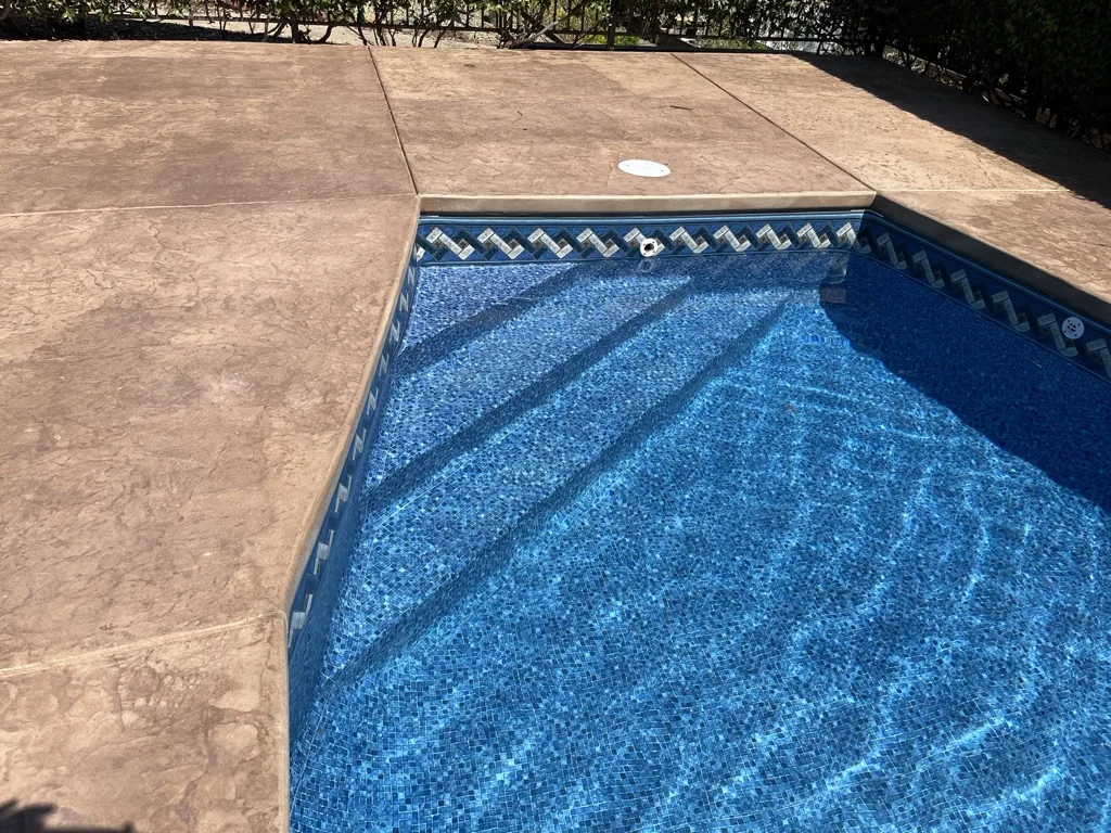 Swimming pool with blue mosaic tile interior and decorative border trim, adjacent to tan concrete deck with dark landscaping