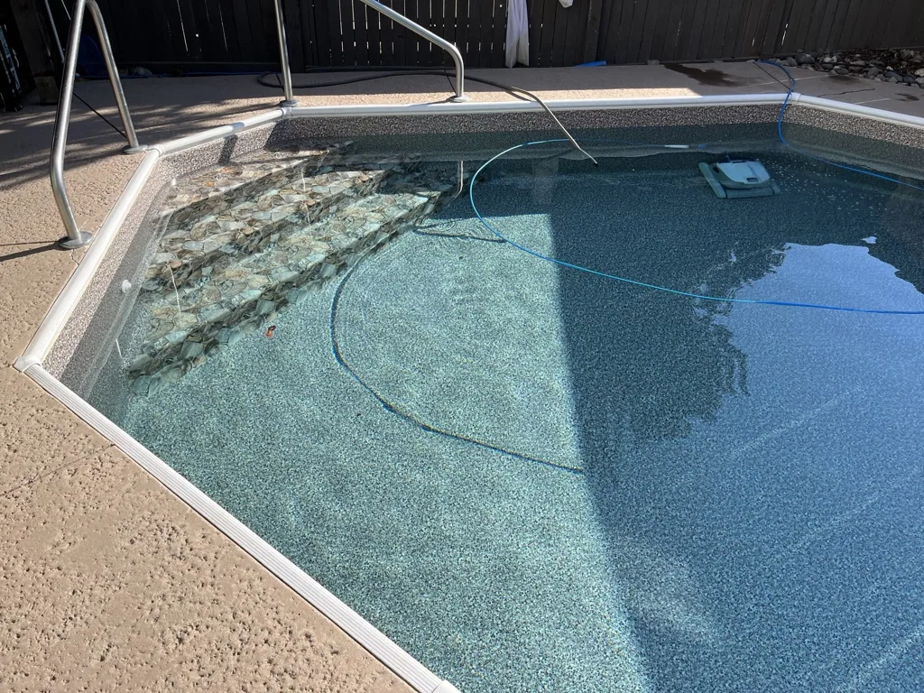 Residential swimming pool with clear turquoise water, metal ladder, and cleaning equipment visible on the deck