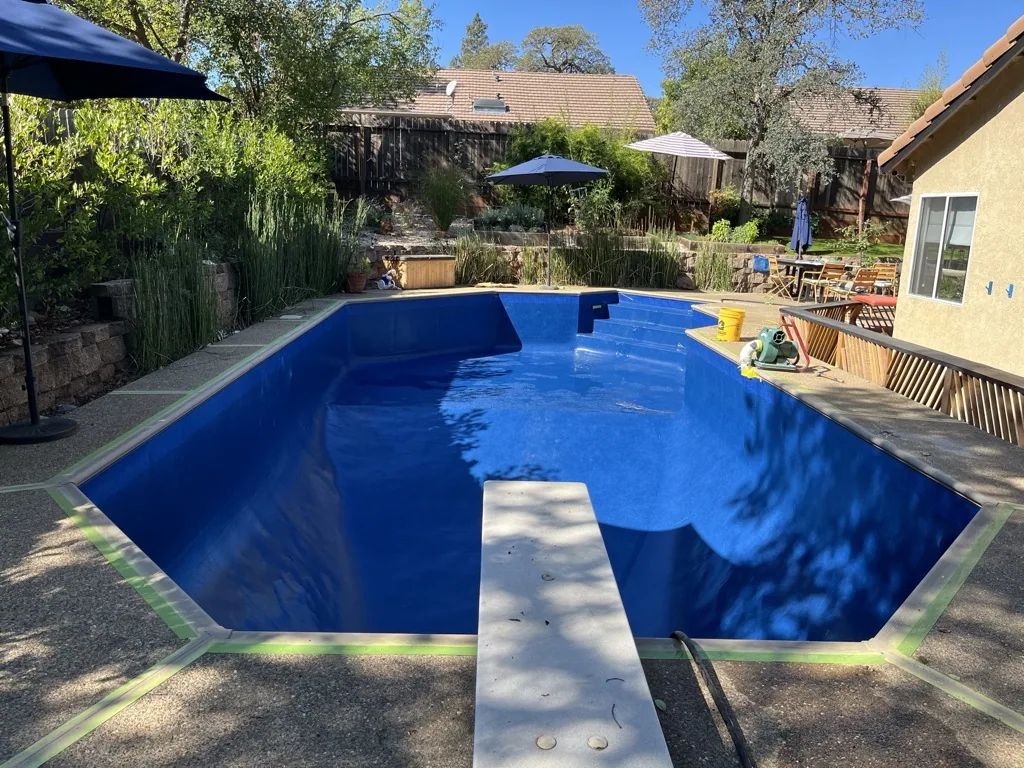 Empty backyard swimming pool with bright blue interior and concrete diving board, surrounded by landscaping and residential buildings