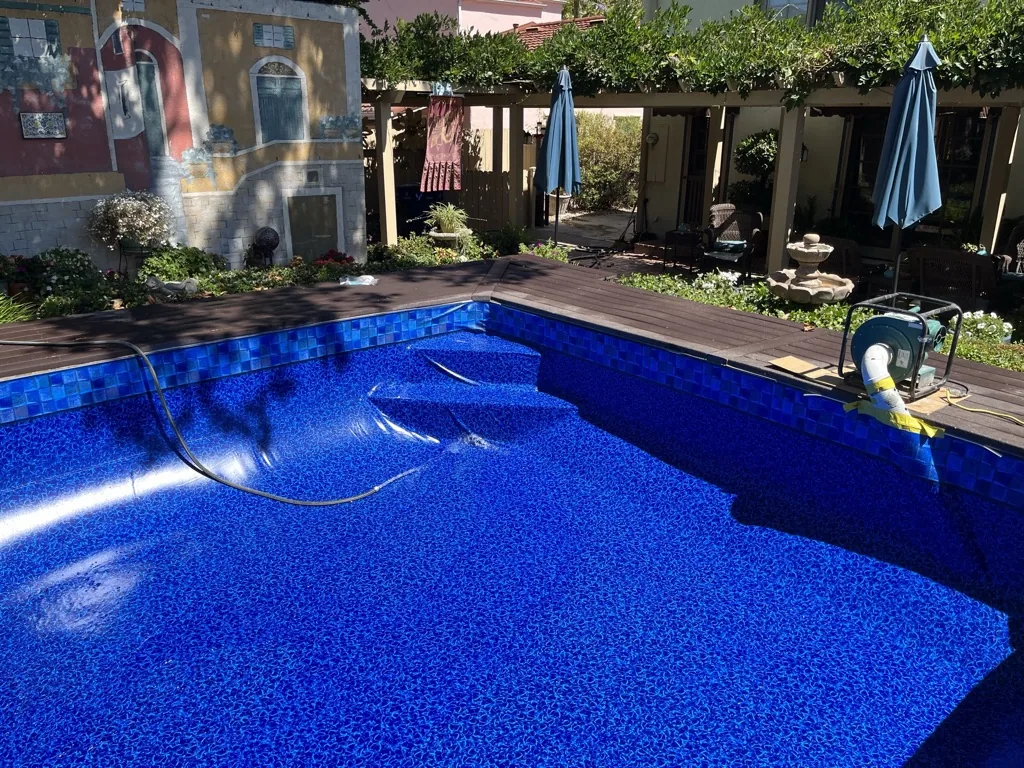Rectangular swimming pool with bright blue water and textured bottom, surrounded by dark wood deck, green pergola, and residential building