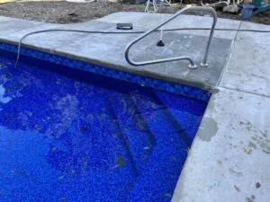 Swimming pool with blue tile interior, concrete deck, metal handrail, and debris on water surface