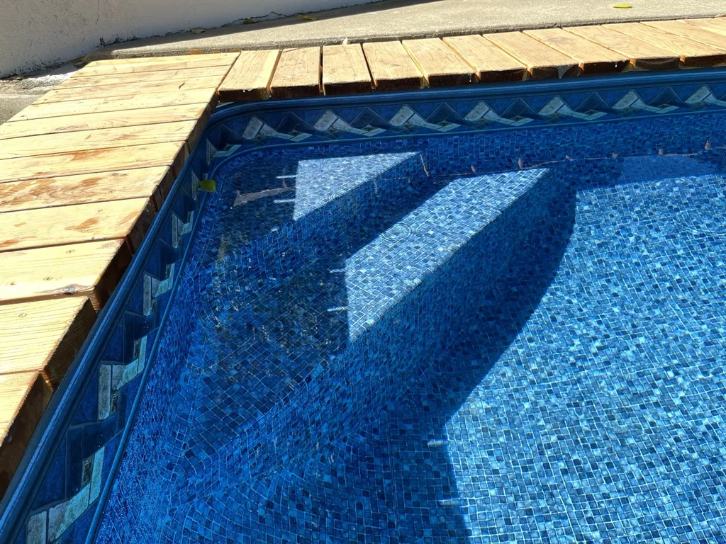Swimming pool with blue mosaic tile interior and wooden deck casting shadows on water surface