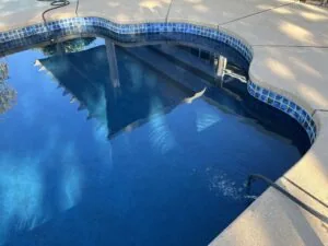 Overhead view of swimming pool with blue tile border and concrete deck, showing underwater steps and sun reflections on water surface.