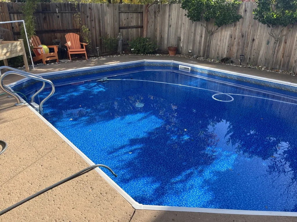 Backyard swimming pool with bright blue water, concrete deck, and wooden fence with two orange Adirondack chairs visible in background.