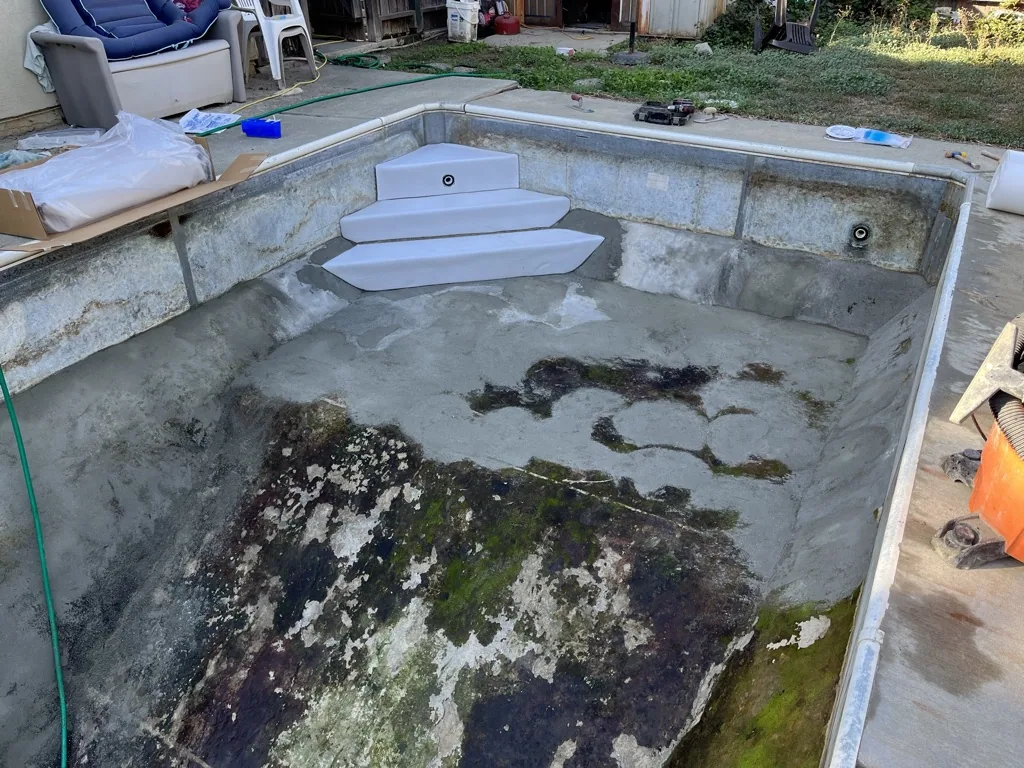 Empty concrete pool with algae and mold stains, white steps, backyard setting with storage items