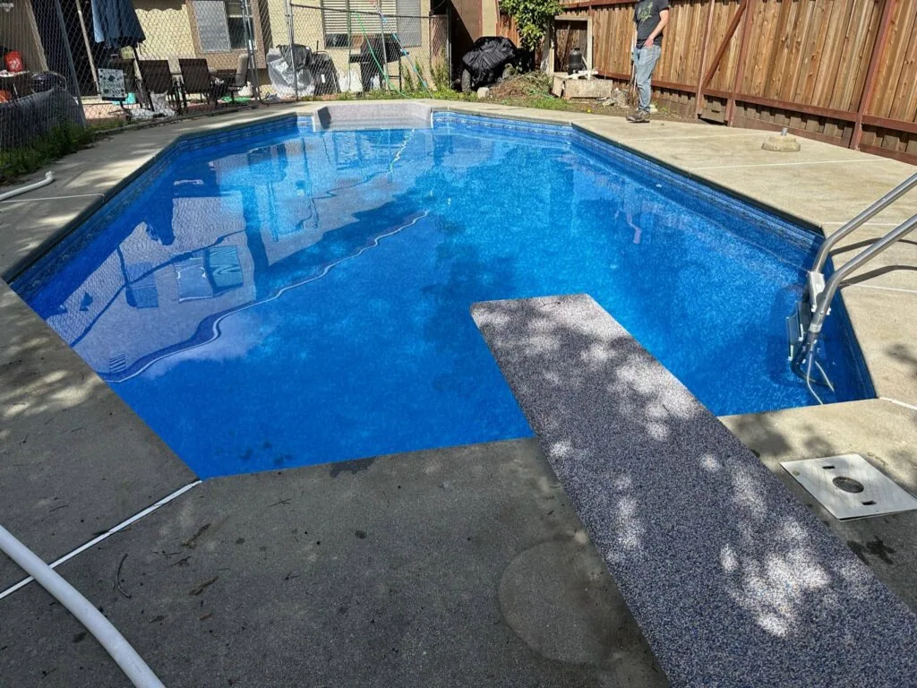 Empty backyard swimming pool with blue interior and attached tanning ledge, surrounded by concrete deck and wooden fence
