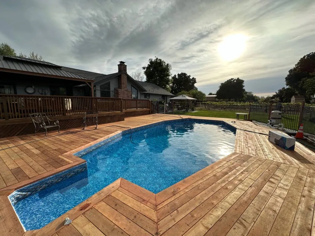 Resort-style swimming pool with blue water surrounded by wooden deck, house with brick chimney, and rural landscape under cloudy sky