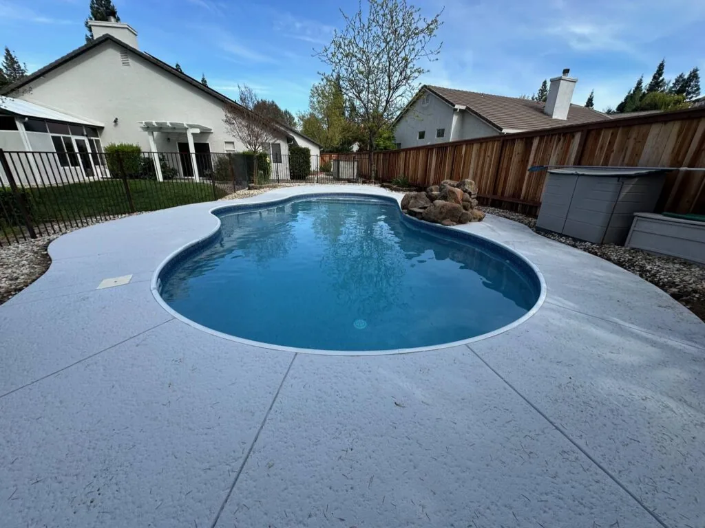 Oval swimming pool with clear blue water surrounded by white concrete decking, with residential houses and wooden fence in background
