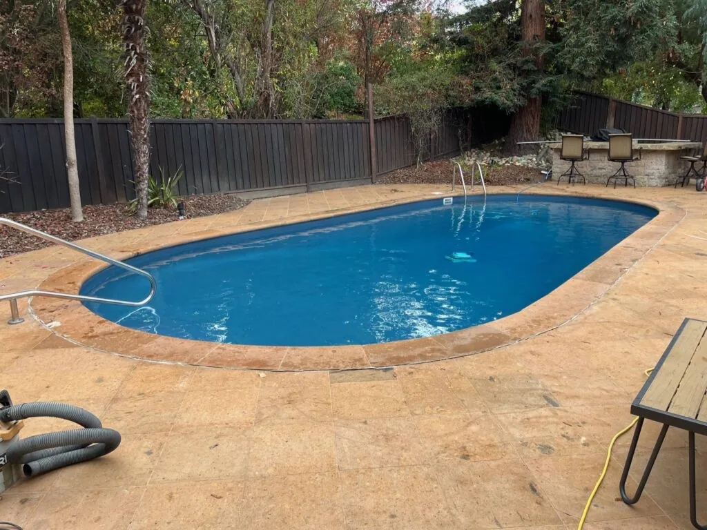 Backyard swimming pool with blue water surrounded by concrete deck, metal fence, and landscaping with tall trees.