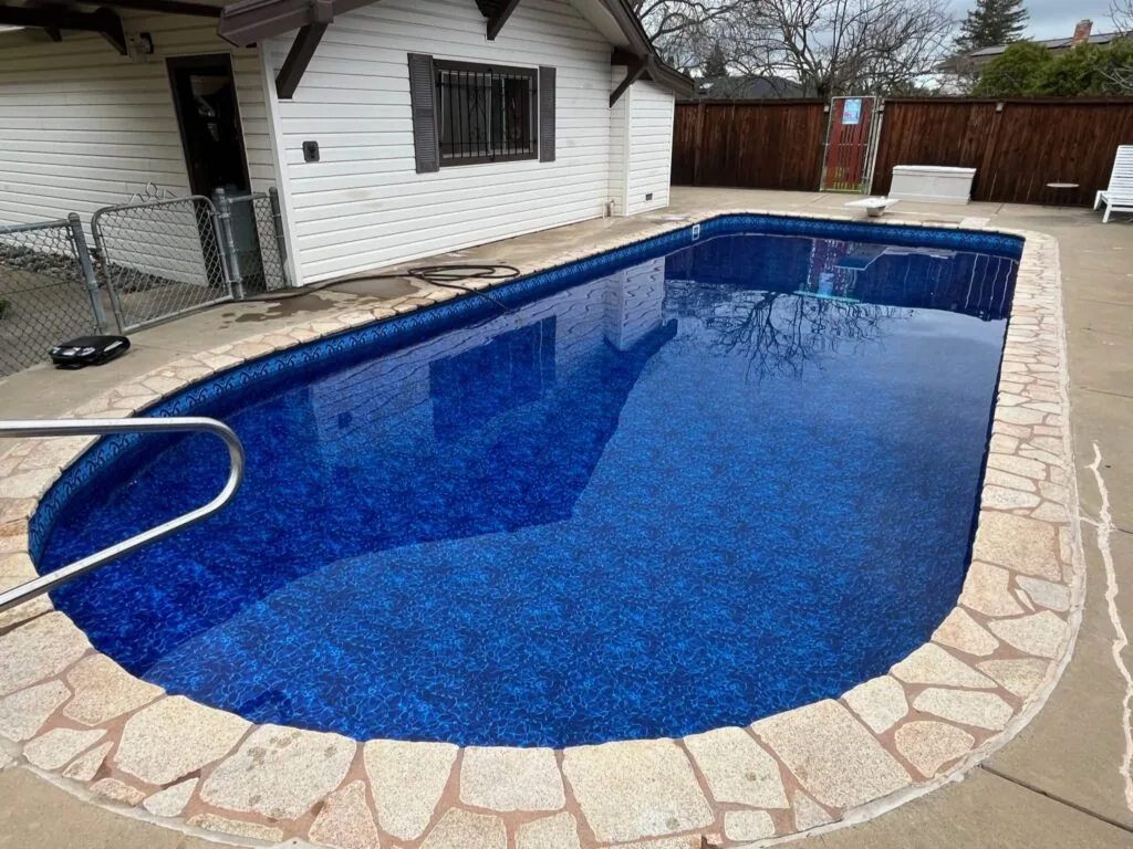 Empty residential swimming pool with blue interior liner surrounded by stone patio, white house with dark roof in background