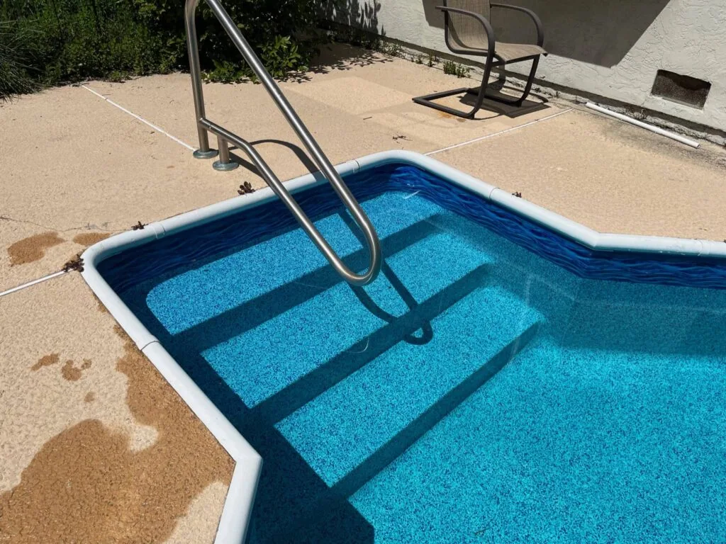 Swimming pool with blue water and submerged steps, metal handrail, and sandy pool deck with lounge chair