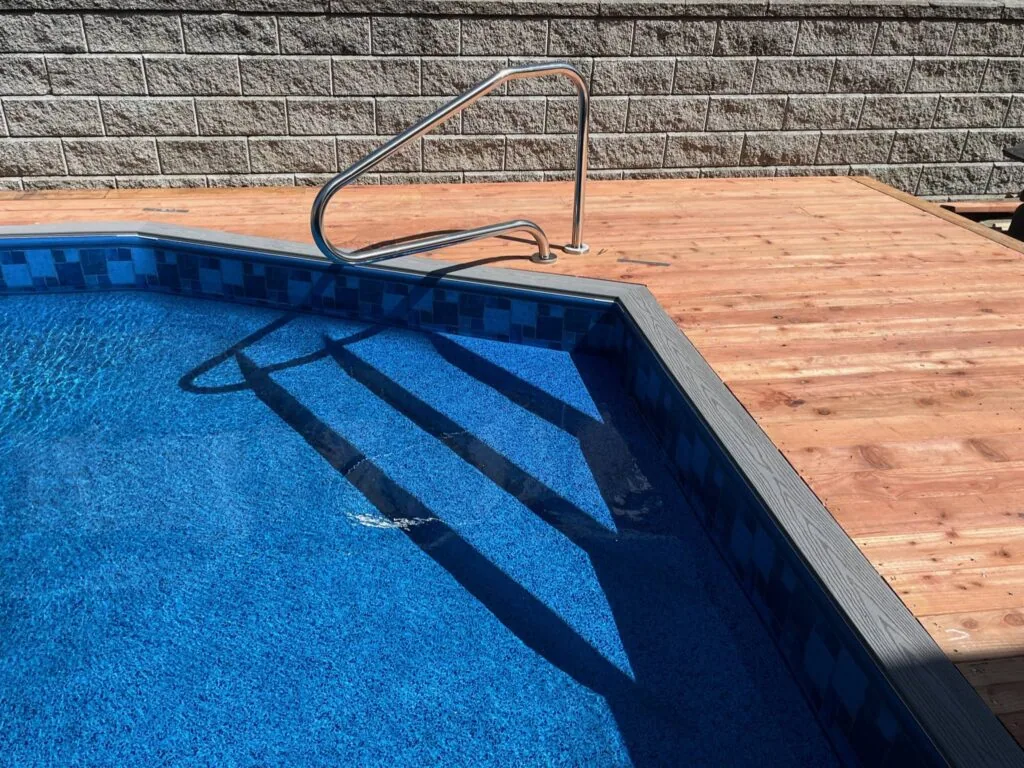 In-ground swimming pool with blue water and ladder beside wooden deck and gray stone wall