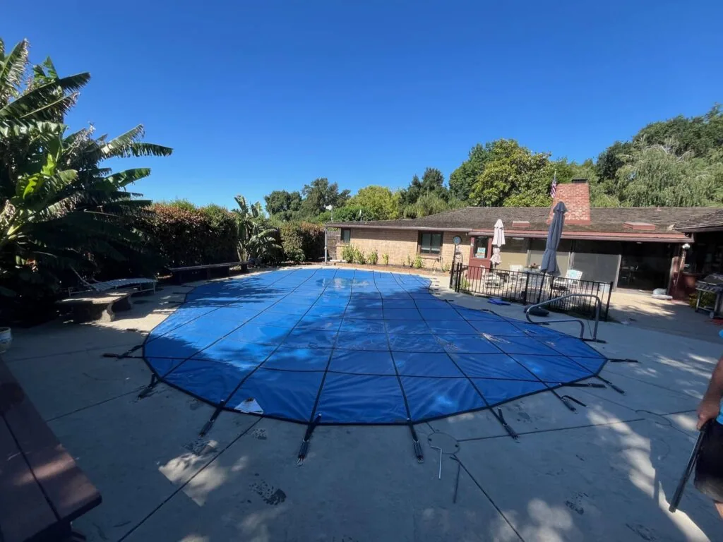 Blue safety cover stretched across rectangular swimming pool with brick pool house and landscaped hillside in background