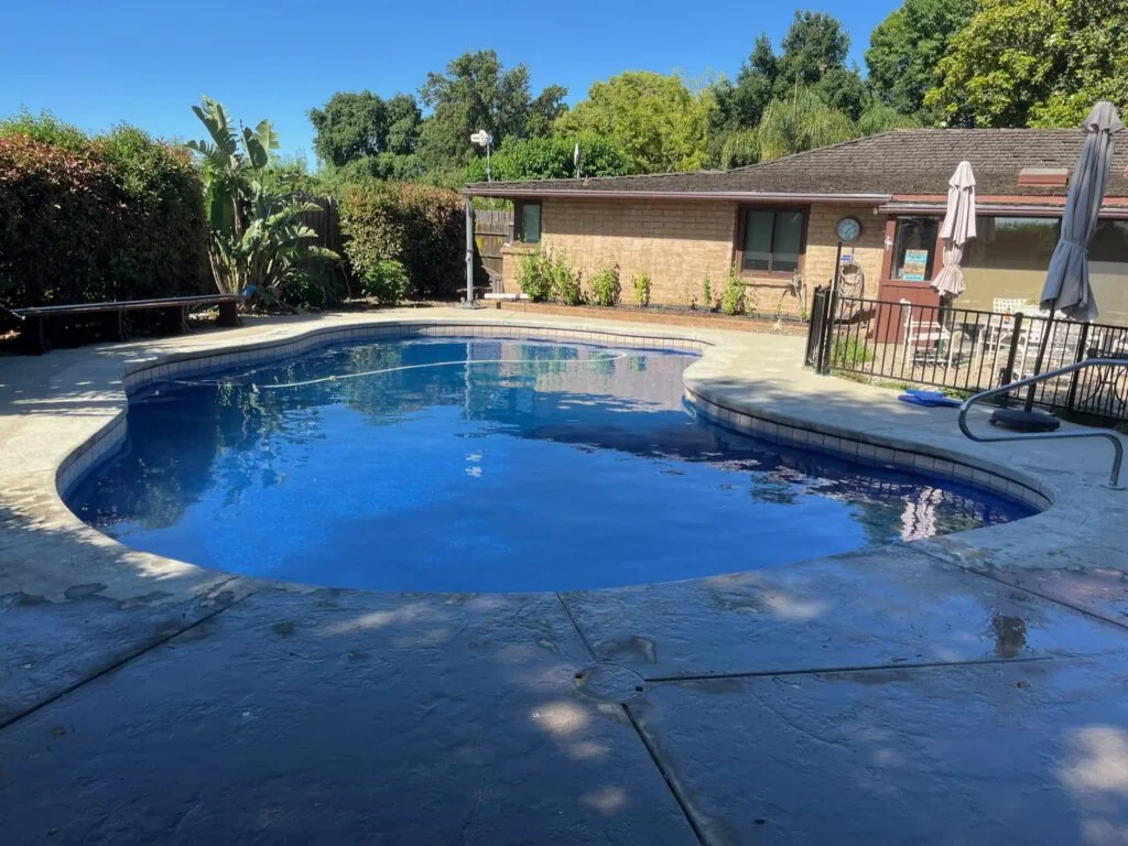 Backyard swimming pool with blue water, concrete patio, brick house, metal fencing, and mature trees under clear sky