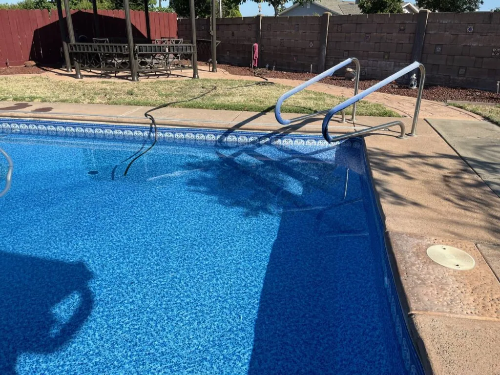 Swimming pool with blue water and metal ladder beside concrete deck, fenced yard in background