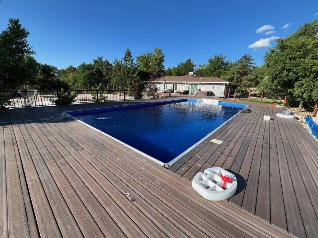 Rectangular swimming pool with blue water and wooden deck surrounding it, with a house and trees in the background under clear blue sky