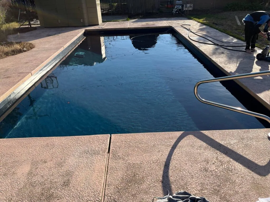 Empty rectangular swimming pool with concrete deck and metal handrail, showing water with dark debris