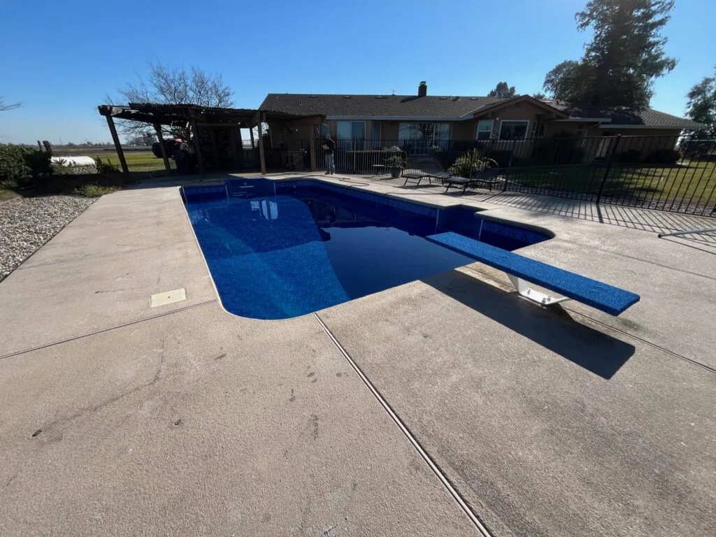 Empty in-ground swimming pool with blue interior and concrete deck, adjacent to residential home with pergola and patio furniture
