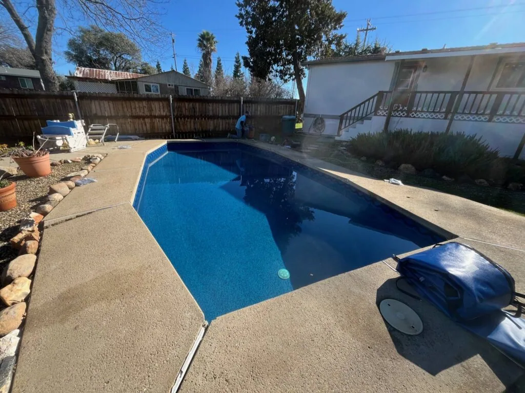 Blue backyard swimming pool with concrete deck, surrounded by fencing and houses with palm trees in background
