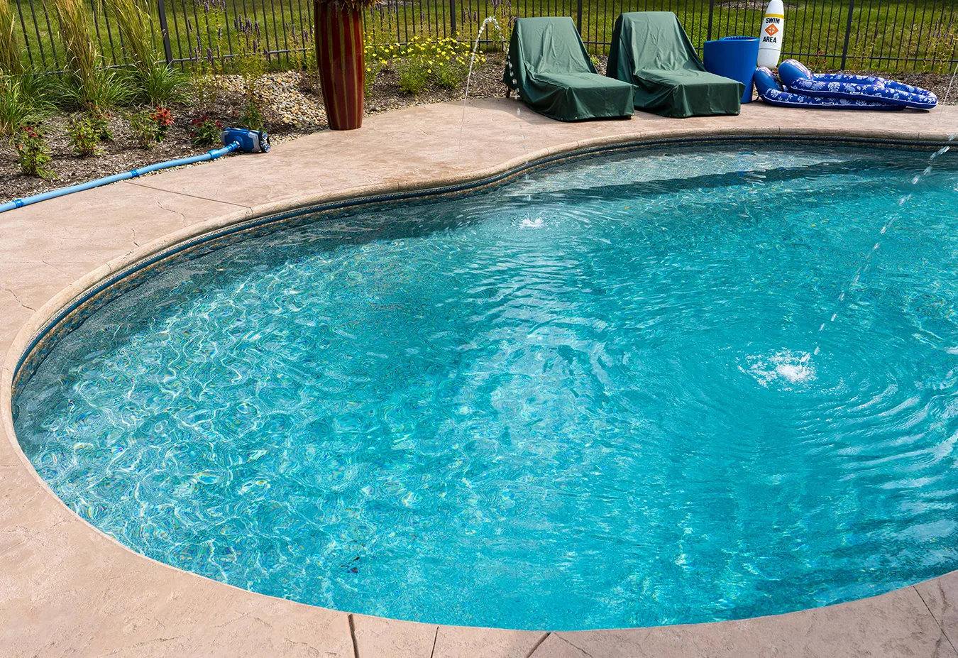 Oval-shaped swimming pool with clear blue water, concrete deck, covered lounge chairs, and fenced landscaping in background