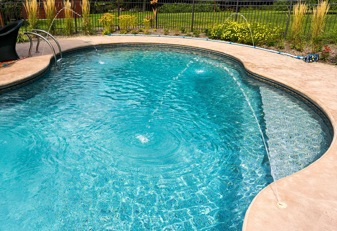 Curved kidney-shaped swimming pool with turquoise water, concrete deck, and fenced yard with landscaping in background