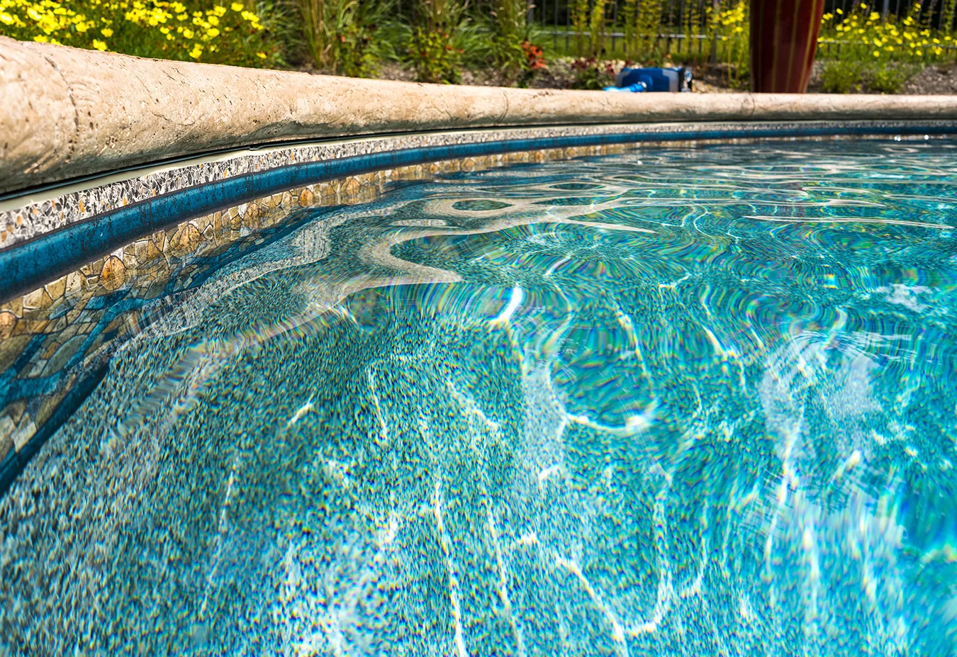 Clear blue water in an in-ground vinyl liner pool with rippling surface and blue-rimmed edge, garden foliage visible in background