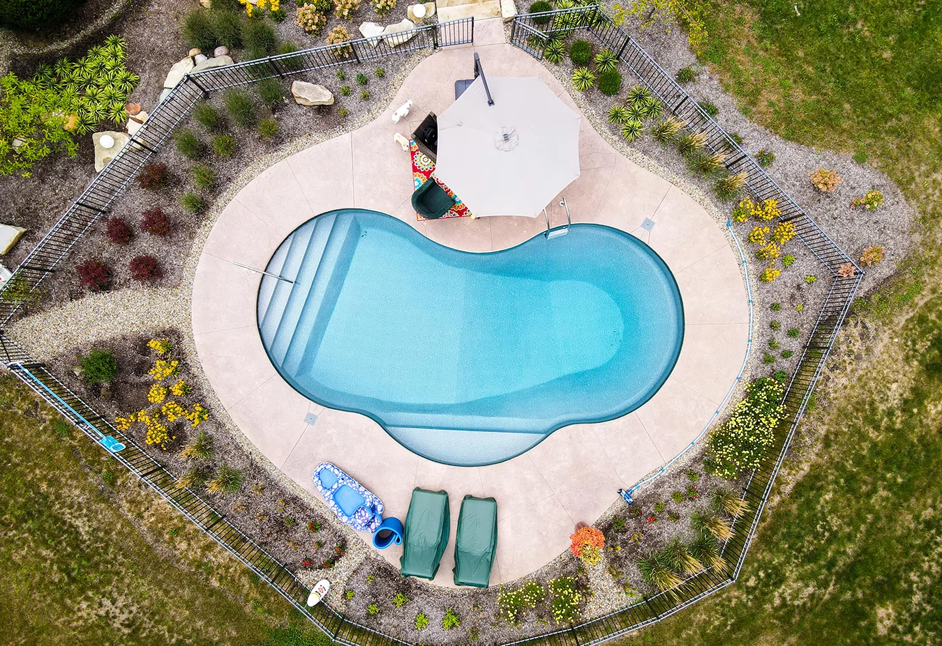 Aerial view of kidney-shaped swimming pool with blue water surrounded by concrete deck, lounge chairs, landscaping, and grass